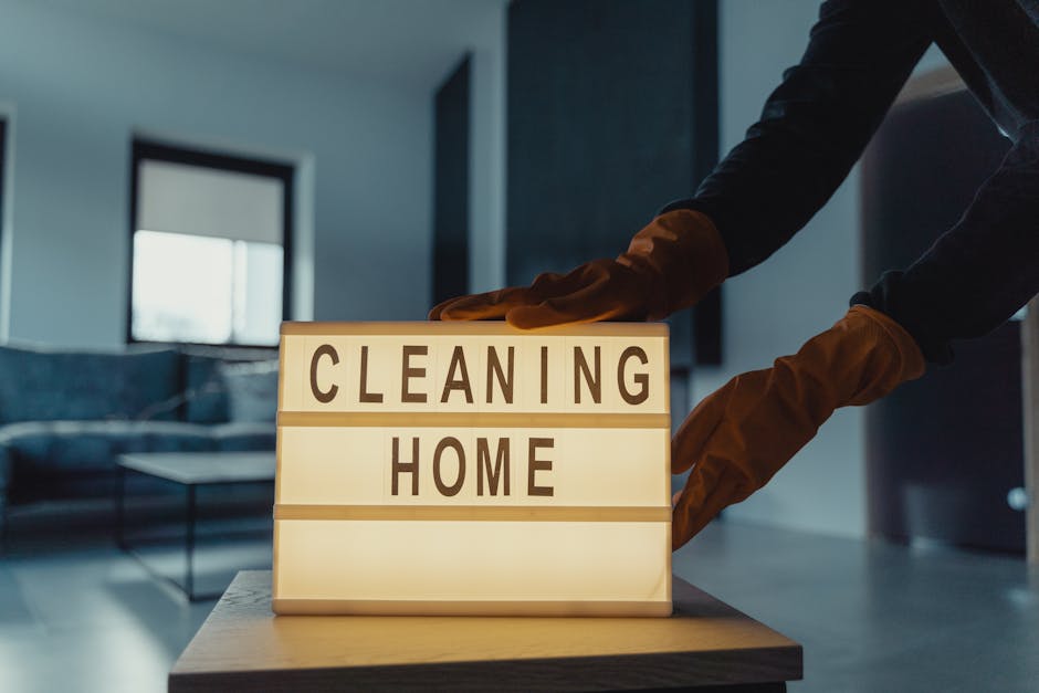 A person wearing orange gloves is placing a lightbox sign with the words 'CLEANING HOME' onto a wooden table in a well-lit modern room. The sign consists of two illuminated panels with black text on a